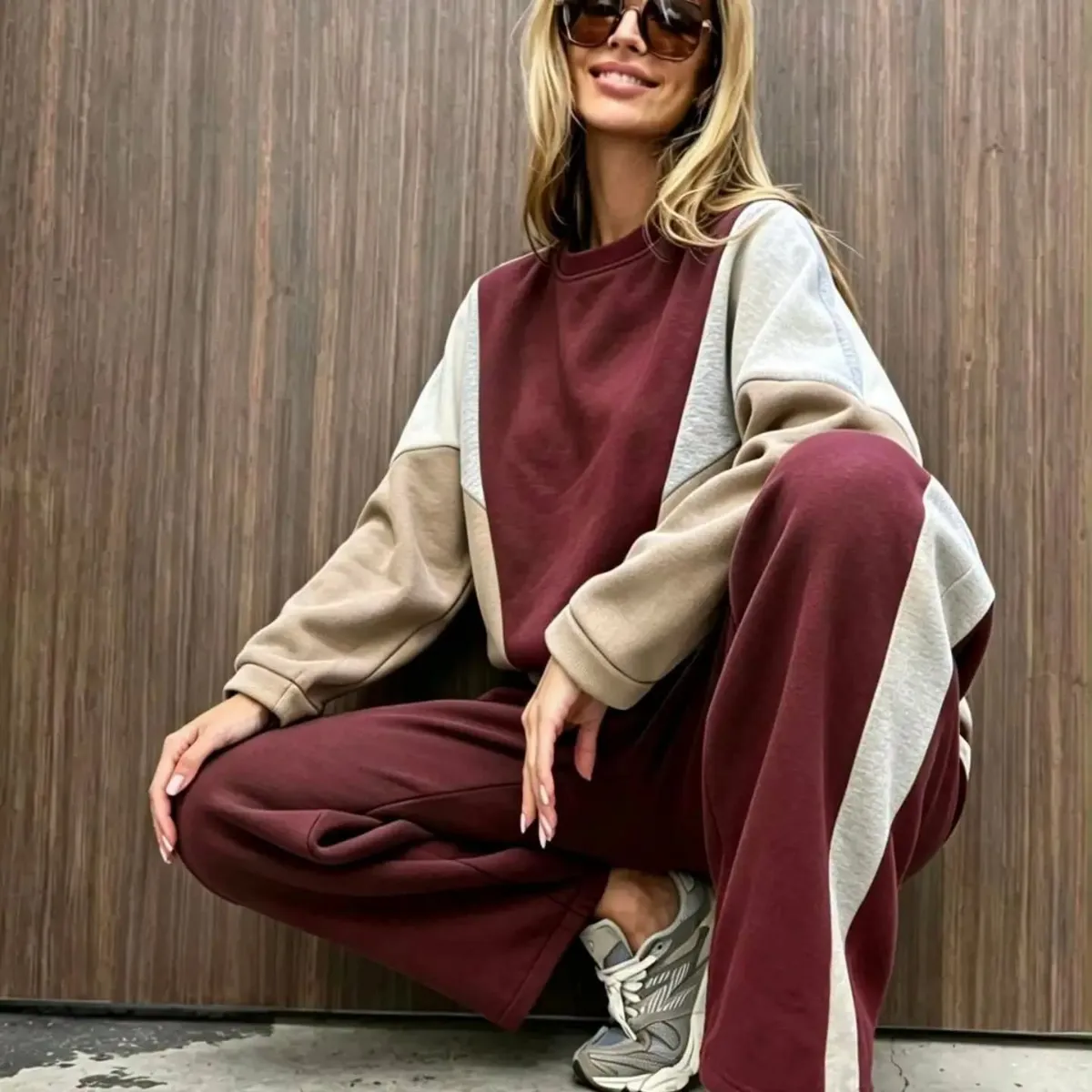 Kvinna i solglasögon sitter på huk framför en trävägg i ett set i vinrött, beige och grått